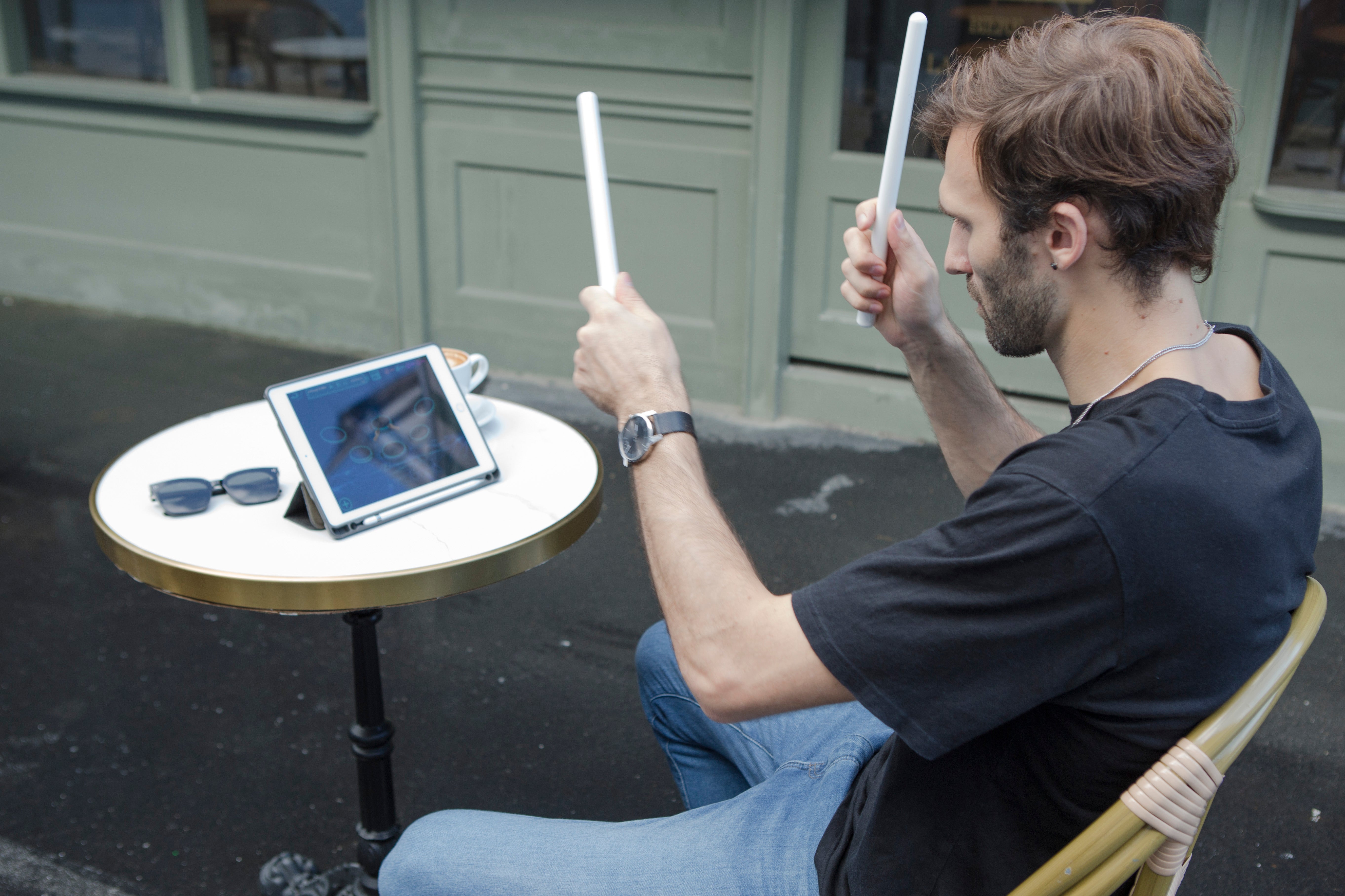 Enjoying a coffee break, a man uses his PocketDrum 2 Max air drumsticks to interact with a music app on his tablet outdoors
